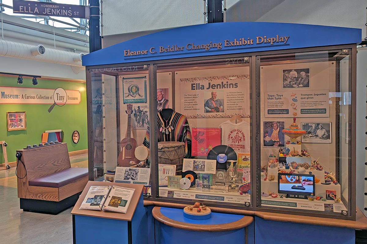 A museum display shows three glass windows containing photos of Ella Jenkins, a poncho and jewelry worn by her, some of her vinyl LP albums, and on the right, her collection of spinning tops.